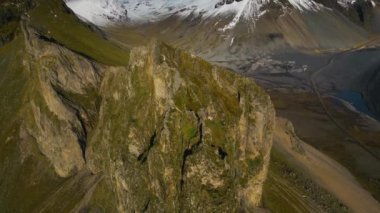 Gündüzleri, bir dron engebeli dağlık araziyi ortaya çıkaran aşağı doğru bir eğim yakalar ve arka planda karla kaplı tepeler belirir.