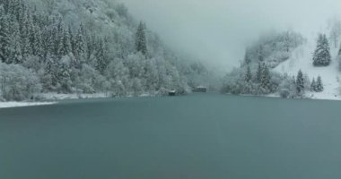 Kar yağışı sırasında bir dron, sık ağaçlarla kaplı bir ormanı keserek gölün üzerinde uçar. Karla kaplı tepeler, kısmen sisle kaplanmış, bulutlu bir gökyüzünün altında arka planda görülebilir.
