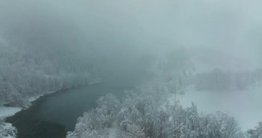Bir dron karda bir gölün üzerinde uçuyor, sık ağaçlarla kaplı bir ormanın içinden geçiyor. Arka planda, karla kaplı tepeler bulutlu bir gökyüzünün altında kısmen sisle kaplıdır.