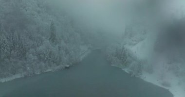 Şiddetli kar yağışı sırasında kısmen sisle kaplanmış, karla kaplı ağaçlar ve dağlarla çevrili bir göl manzarası.