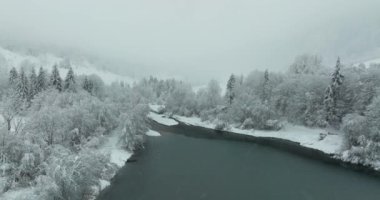 Bir dron güzel bir kış manzarasının üzerinde uçar, karla kaplı ağaçlarla çevrili sakin göl manzarası sunar, uzak tepeler ve dağlar kısmen gri gökyüzünün altında sisle kaplıdır.