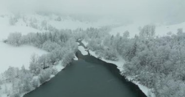 Kış manzarası üzerinde uçan bir dron, karla kaplı ağaçlarla çevrili bir gölün üzerinden geçerken, bulutlu bir gökyüzünün altında kısmen gizlenmiş bir dağ zeminine doğru uçuyor.
