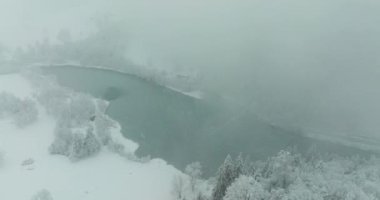 Donmuş bir gölün üzerinde uçan bir dronun karla kaplı sık ormanlarla çevrili bir görüntüsü. Yoğun kar yağışı sırasında etrafındaki tepeler ve kış manzaraları kısmen sisle kaplıdır.
