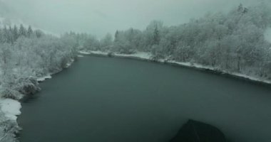 Karla kaplı bir çam ormanıyla çevrili donmuş bir gölün üzerinde bir dron süzülüyor. Görünür kar çizgileri bulutlu gökyüzünün altında dağıldı.