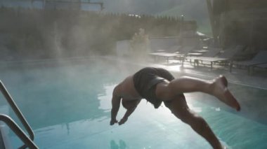 Arka plan görüntüsü, kaplıcadaki sisli bir havuza yavaş çekimde dalan bir adamı yakalar. Havuz şezlonglar ve doğa ile çevrili, sakin ve lüks bir atmosfer yaratıyor.