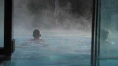 Dış kaplıcada yüzen bir adamla bir kadının arka plan görüntüsü. Ön plandaki cam kapı yavaşça kapanırken havuzdan buhar yükseliyor.