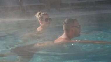 Buharlı bir kaplıca havuzunda şezlonglu arka planda yüzerken bir kadın arkadaşına gülümsüyor.