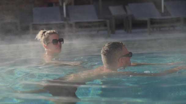 ハッピーカップルのスローモーションショットは 日光の下でスチームアウトドアスパプールで泳いでいる間チャットをしています — ストック動画
