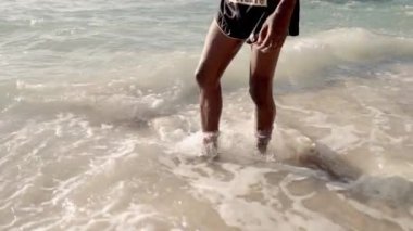 Bir Muay Thai boksörünün yakın plan çekimi kumlu kıyılarda dimdik ayakta duruyor, bacaklarını okyanus dalgalarının ritmik dansının ortasında