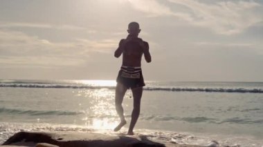 Bir Muay Thai dövüşçüsü kumlu bir sahilde, sakin bir okyanus zeminine karşı güçlü tekme ve yumruk pratiği yapar.