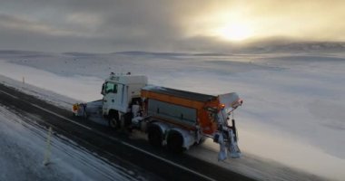 Buzlu bir yolu temizlerken bir dron kar küreme aracının üzerinden uçar. Güneş kış boyunca İzlanda 'nın karla kaplı topraklarına sıcak bir parıltı saçar.