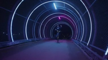 Becerikli bir BMX binicisi gece vakti zincirleme tünelin ışıkları altında etkileyici manüel numaralar sergiliyor.