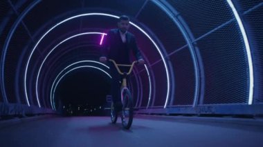 Bir BMX sürücüsü, gece vakti neon ışıklarıyla bir zincir-bağlantı tünelinde ustalıkla gezinerek burun kullanma kılavuzlarını ustalıkla yapar.