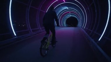Becerikli bir BMX sürücüsü, neon ışıklarla donatılmış bir yaya köprüsünde 360 derecelik bir dönüşle göz kamaştırıyor. Dinamik bir arka görüş açısından çekiliyor.