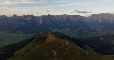 İHA 'dan gelen bir hava görüntüsü, açık mavi gökyüzünün altındaki yemyeşil bir yamaçtan geçerek, uzak bir dağ sırasına yavaşça yaklaşır.