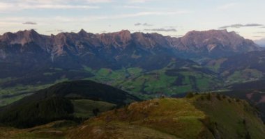 Uçan bir dronun engebeli bir dağın tepesinden yemyeşil yamaçlar ve arazilerle dolu, mavi gökyüzünün altındaki uzak dağ sırasının zeminine kurulmuş görüntüsü