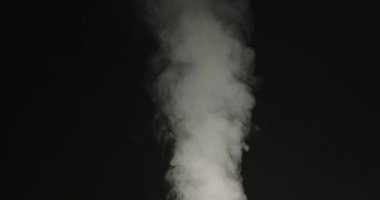 Yoğun beyaz duman siyah arkaplana karşı girdap halinde yükselir ve havaya karışmadan önce karmaşık şekiller oluşturur.