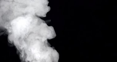 Kalın beyaz duman siyah arka plana doğru dönen bir hareketle yükselir. Parlak ışık, karmaşık formunu ve şablonlarını yavaş çekimde vurgular.