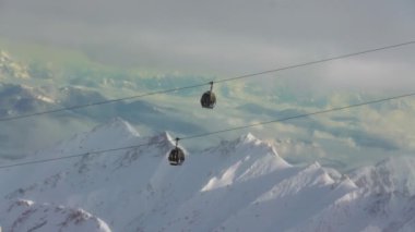 Çelik bir kabloda asılı iki teleferiğin karla kaplı dağlık bir arazide uçuştuğu bir hava görüntüsü. Gökyüzü parçalı bulutlu, güneş dağlarda gölgeler ve gölgeler bırakıyor.