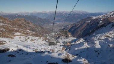 Açık mavi gökyüzünün altında karlı bir dağ manzarasında yolcuları taşıyan bir kayak asansörü. Arka plan, uzak dağ ve vadilerin parlak güneş ışığıyla yıkanmış panoramik bir görüntüsünü ortaya çıkarıyor.