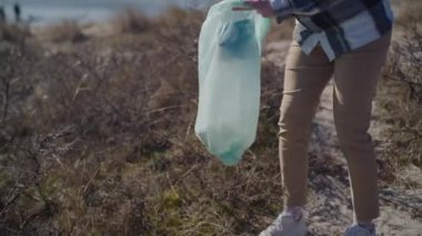 A tracking shot of a woman cleaning up trash scattered along dried grass