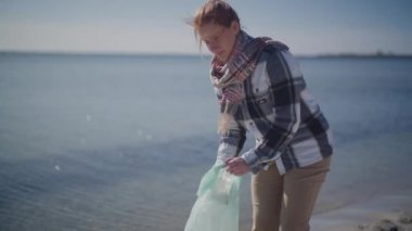 A tracking shot of a woman collecting trash along the shore during daytime