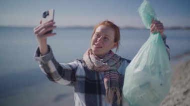 A handheld shot zooming out from a woman standing on a serene beach, engaged in a video call while holding a trash bag