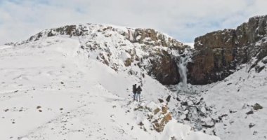 Bir dağ yamacında, nefes kesici bir şelale ve engebeli bir kar manzarasıyla çevrili iki insanın hava görüntüsü.