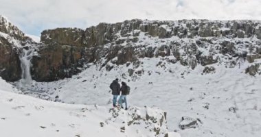 Bir dron karlı bir uçurumun tepesinde duran iki insanı yakalıyor, uzak bir şelalenin nefes kesici manzarasına hayret ediyor.