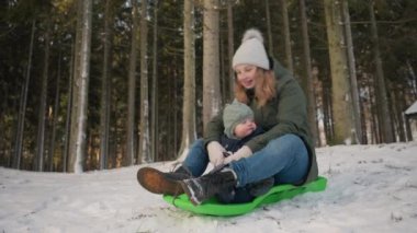 Yükselen ağaçların sükunetiyle çevrili karlı bir ormanda süzülen bir anne ve bebeğin iz sürme görüntüsü.