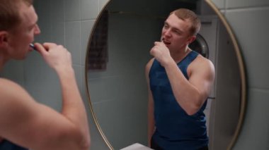 Bir adam banyo lavabosunun yanında durup aynaya bakarken dişlerini fırçalıyor ve günlük rutininin bir parçası olarak eksiksiz bir temizlik sağlıyor.