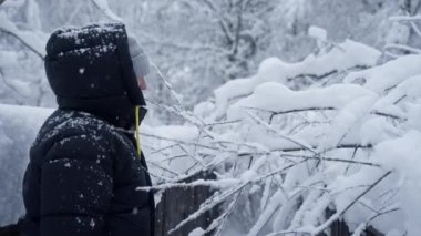 Siyah kabarık ceketli bir adam, kar dolu dalları özenle çeker ve temizler. Kış ayazının dingin bir zemin üzerindeki keskin güzelliğini gözler önüne serer.