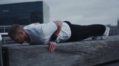 Dışarıdaki tahta kirişte şınav çeken bir adam. Beyaz tişört, siyah pantolon ve beyaz spor ayakkabı giyiyor. Arkaplanda bulutlu bir gökyüzü ve şehir mimarisi yer alıyor.