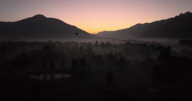 Güneşin doğduğu gökyüzünün altındaki görkemli dağların çerçevelediği siluetli ağaçlardan oluşan bir tarlada sisli bir manzara beliriyor.