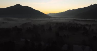 Bir dron, gün doğumunda siluetli dağlara yuva yapmış sakin ve sisli bir kasabayı ortaya çıkarmak için yükselir ve sakin bir sabah manzarası yaratır.