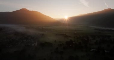 Güneşin doğduğu sisli bir kasabanın üzerine güneş ışığı gibi iniyor, dingin bir sabah sisinin ortasında ruhani ışık ve gölgeler bırakıyor.