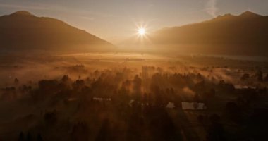 Panoramik bir çekim sisli bir ormanda uzak siluetli dağların arasından geçiyor. Her ikisi de güneşin sıcacık parıltısıyla yıkanıyor.
