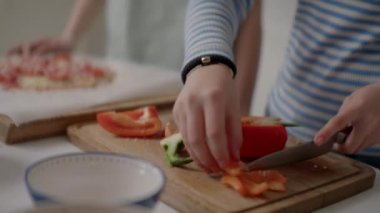 Yakın plan çekimler, ahşap kesme tahtasındaki taze dolma biberleri ustalıkla dilimlemek, ev yapımı pizzaya canlı malzemeler hazırlamak. Arka planda, malzemelerle dolu bir pizza pişmeyi bekliyor.