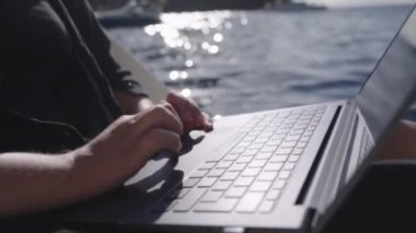 Laptop klavyesine yazı yazan bir kadının yakın çekimi. Dizüstü bilgisayar, arka planda bulanık bir deniz manzarasıyla onun kucağında duruyor..
