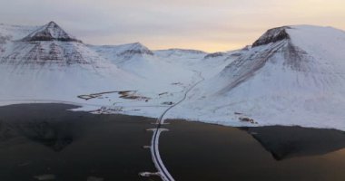 Görkemli karlı dağlarla çerçevelenmiş sakin bir göl üzerindeki bir yolun manzarası, doğanın kış manzarasının dingin güzelliğini gözler önüne seriyor.
