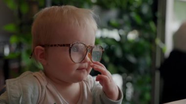 Yuvarlak gözlük takan sevimli bir bebeğin yakın çekimi. Çocuk, gözlüğü kısaca ayarlayan bir yetişkinin eliyle etkileşime giriyor..