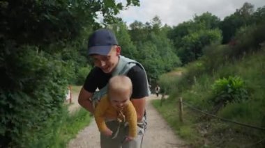 Genç bir çocuk bir bebekle oynar. Çocuk koşuyor, bebeği önünde tutuyor. Bebek kahverengi tulum giyiyor ve sarı saçları var. Çamurlu bir yolda yeşilliklerle çevrili.