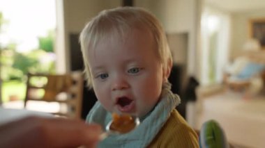 Yakın plan çekimde kaşıkta bebek mamasıyla beslenen bir bebek görülüyor. Bir yetişkinin eli kaşığı tutar, arka planda hafifçe iç kısımları bulanık olur.