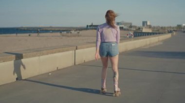 Bir kadın plajın yakınındaki kaldırımlı bir patikada paten kayıyor. Kadının saçları rüzgarda uçuşuyor ve kameradan uzaklaşıyor.