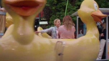 İki kadın mutlu bir şekilde ördek şeklinde bir atlıkarıncaya biniyor. Atlıkarınca dönerken kollarını kaldırıp gülüyorlar. Arka planda panayır tezgahları ve dekoratif duvarlar görülebilir.