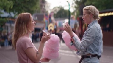 Elinde pamuk şeker olan iki genç kadın bir panayırda karşı karşıya duruyor. Sağdaki kadın parmaklarını uzatarak arkadaşına bir parça pamuk şeker sunuyor.