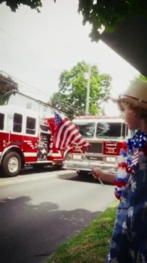 Küçük bir çocuk, bir Amerikan bayrağını tutuyor ve bir itfaiye aracının yanından geçerken coşkuyla el sallıyor.