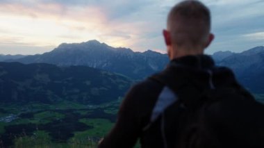 Bir adam kolunu uzatır ve batan güneşin aydınlattığı kayalık dağları gösterir.