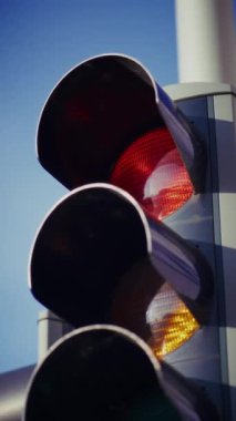 Yakından çekilen bir fotoğraf, parlak, modern bir kılıfla korunan ve doğrudan güneş ışığından korunan bir trafik ışığını yakalar, açık mavi gökyüzüne karşı koyu renklerle keskin bir tezat oluşturur.