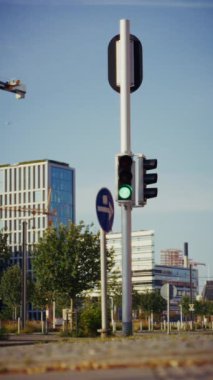 Bir trafik sinyali yeşilden kırmızıya geçerken, araçların durması gerektiğini belirtirken, yakınlardaki bir kutuptaki mobese kamerası da yeşil ve modern binalarla trafiği izliyor.
